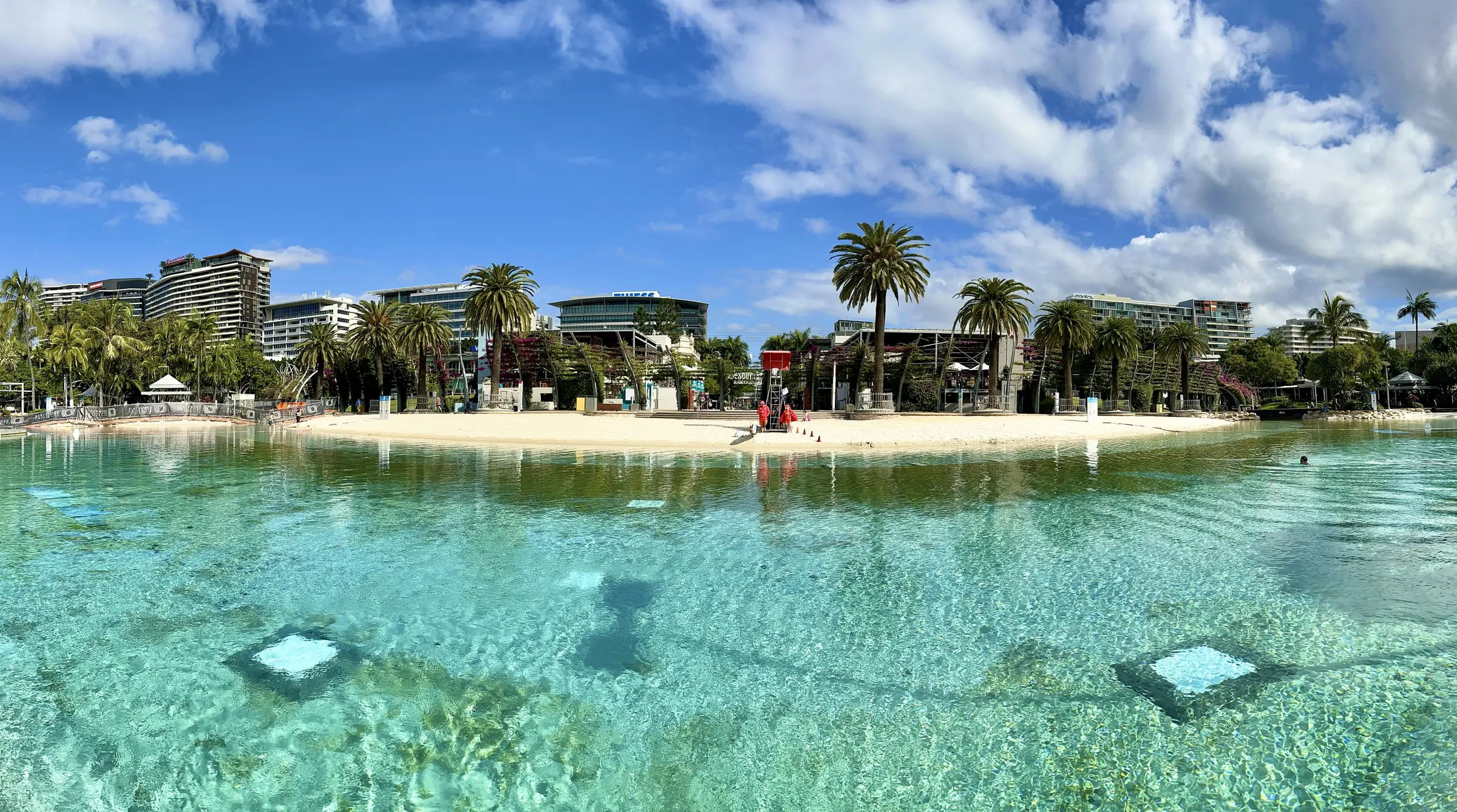 Streets Beach at South Bank: a clear turquoise lagoon over pale sand, palms and low buildings beyond, under a blue sky full of cloud