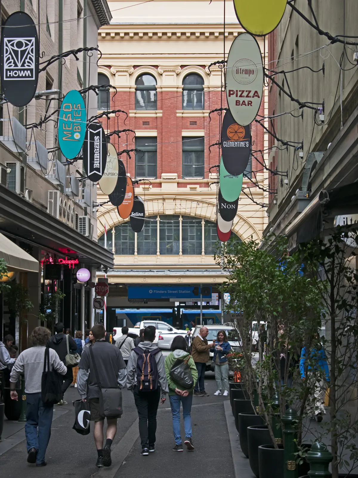 A narrow cafe lane hung with round shop signs, people walking down it toward a red-brick and cream building at its end
