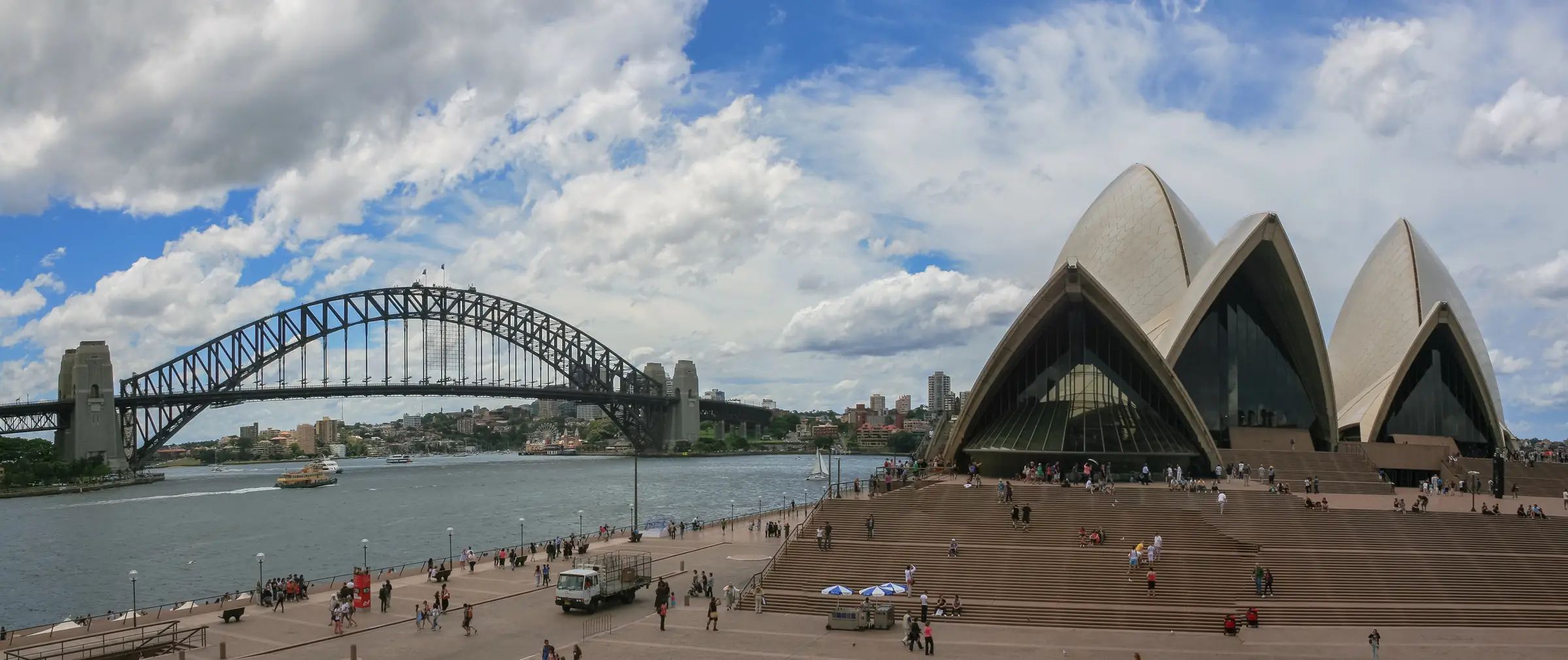 People walking on the Opera House forecourt and steps, with the Harbour Bridge across the water